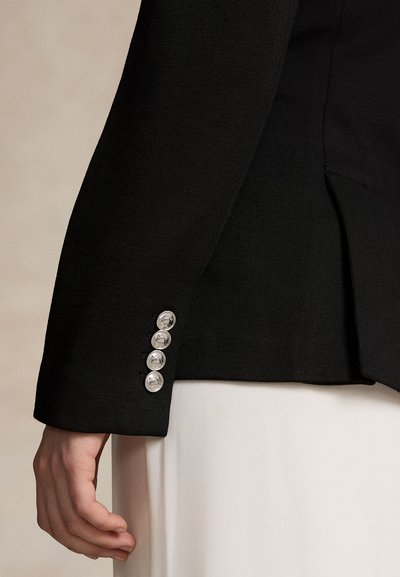 Prim-plan asupra unei mâini lângă manseta unui sacou negru cu patru butoane argintii gravate cu cai, purtat peste o țesătură albă.