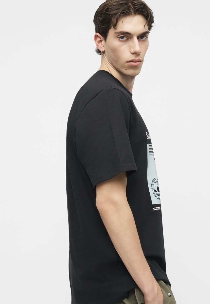 Junger Mann mit kurzen dunklen Haaren, der ein schwarzes Grafik-T-Shirt trägt, seitlich mit neutralem Gesichtsausdruck vor einem einfarbigen weißen Hintergrund.