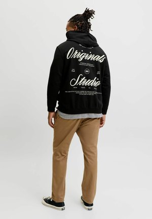 Persoon met donker gevlochten haar die een zwarte hoodie met witte tekst "Originals Studio" op de rug draagt, beige broek en zwarte sneakers, staand met de rug naar ons toe.