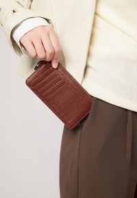 Brauner strukturierten Leder Geldbeutel mit mehreren Kartenfächern, der teilweise aus einer Hosentasche heraus gehalten wird. Glattes Design mit einem Reißverschluss.