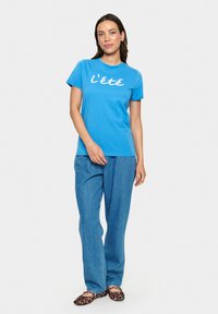 T-shirt en coton bleu avec le texte blanc "l'été", associé à un pantalon en denim bleu clair et des ballerines à imprimé léopard. Modèle se tenant devant un fond uni.
