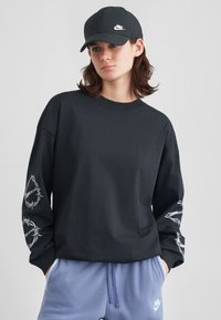 Mladý človek v čiernej šiltovke Nike a čiernom tričku s dlhým rukávom s bielou grafikou na rukávoch, v kombinácii s modrými teplákmi Nike.