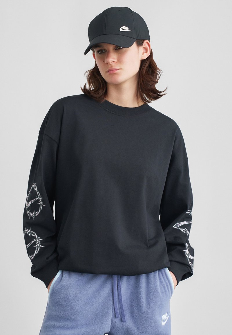 Mladý človek v čiernej šiltovke Nike a čiernom tričku s dlhým rukávom s bielou grafikou na rukávoch, v kombinácii s modrými teplákmi Nike.