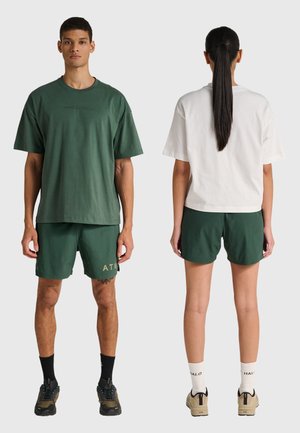 Mand står med fronten mod kameraet iført grøn oversized T-shirt og shorts, kvinde står med ryggen mod kameraet iført hvid oversized T-shirt og grønne shorts med hestehale.