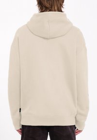 Hoodie beige avec une texture douce, doté d'une capuche et de poignets côtelés. Le dos ne présente aucune impression ni motif, avec une petite étiquette logo au niveau de l'ourlet.