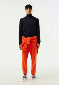 Marinblå turtleneck-tröja och orange joggerbyxor, med en tröja knuten runt midjan. Modell som bär färgglada sneakers, stående framför en vit bakgrund.