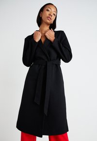Long black coat made of textured fabric, featuring a wrap design, wide collar, and a belted waist. Seen with red pants underneath.