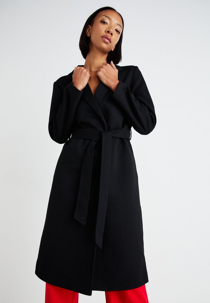 Lungo cappotto nero realizzato in tessuto testurizzato, con un design avvolgente, colletto ampio e vita cintata. Indossato con pantaloni rossi sotto.