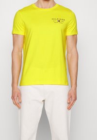 Muž oblečený v jasně žlutém tričku s krátkým rukávem a logem "Hilfiger" na hrudi, s bílými kalhotami, stojící na jednobarevném pozadí.