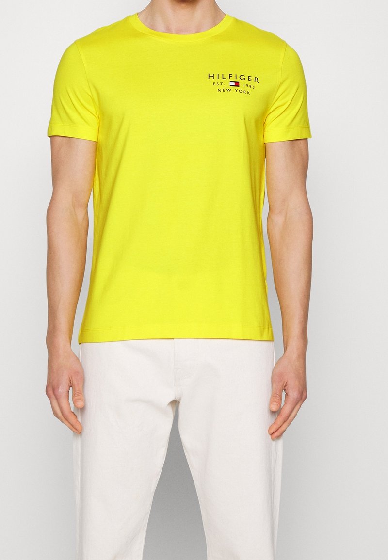 Muž oblečený v jasně žlutém tričku s krátkým rukávem a logem "Hilfiger" na hrudi, s bílými kalhotami, stojící na jednobarevném pozadí.
