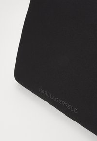 Czarny dodatek z tkaniny o gładkiej fakturze, z wytłoczonym logo "KARL LAGERFELD" w delikatnym matowym wykończeniu. Prostokątny kształt.