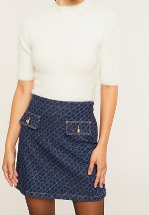 Femme portant un haut court blanc duveteux et une jupe bleu marine à motifs taille haute avec des poches boutonnées à l'avant.