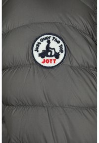 Veste doudoune grise avec texture matelassée. Elle présente un patch brodé rond avec "Just Over The Top" et un design de scooter, ainsi que "JOTT" en rouge.