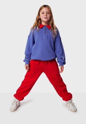 Pull bleu avec col rouge, doté de trois boutons ; associé à un pantalon de jogging rouge. Le matériau semble doux, adapté à un usage décontracté.