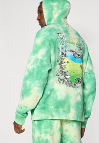 Φαρδιά φούτερ με κουκούλα σε πράσινο tie-dye, με σχέδιο γραφικών στην πλάτη που περιλαμβάνει λουλουδένια στοιχεία, καρτουνίστικες εικόνες και ένα τοπίο, φτιαγμένο από απαλό ύφασμα.