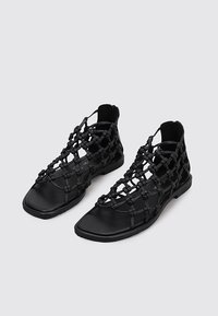 Sandalias planas negras con punteras cuadradas y un intrincado diseño de tiras anudadas que cubren el pie y el tobillo, mostradas sobre un fondo liso.