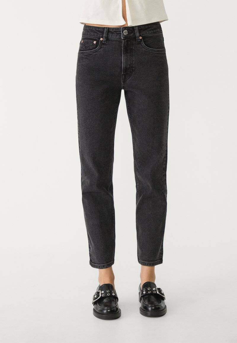 Persona che indossa jeans neri a vita alta abbinati a mocassini in pelle nera con fibbie e occhielli argentati, su uno sfondo semplice.