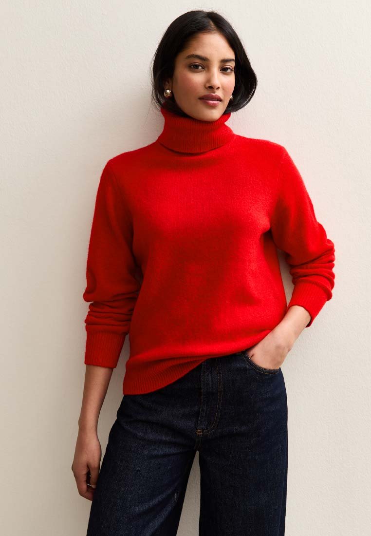 Maglione dolcevita rosso in tessuto morbido a maglia con polsini e orlo a coste, abbinato a jeans blu scuro. Il modello è in piedi davanti a uno sfondo neutro.
