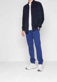 Marinblå kordflorbutton-up skjorta, vit t-shirt med rund hals, blå byxor och vita sneakers med svarta detaljer. Synlig bröstficka på skjortan.