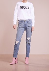 Sweatshirt blanc avec logo "HUGO" noir, jean bleu clair délavé avec déchirures, et bottes à talons hauts roses avec accents métalliques.