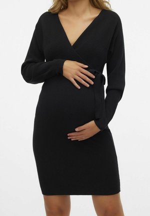Kobieta w ciąży w czarnej, prążkowanej, dzianinowej sukience kopertowej z długim rękawem i wiązaniem z boku, delikatnie obejmująca brzuch dłonie.