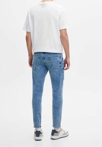 Hombre de pie, de espaldas, vistiendo una camiseta blanca lisa, jeans ajustados de color azul claro, calcetines blancos y zapatillas en blanco y gris.