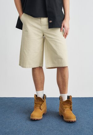 Persona con pantalones cortos beige hasta la rodilla, calcetas blancas y botas de trabajo color tan, de pie sobre una alfombra azul con las manos en los bolsillos.