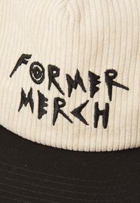 Kremfarget kordfløyelslue med svart brodert tekst "FORMER MERCH." Har en strukturert svart skjerm og rillet tekstur på toppen.