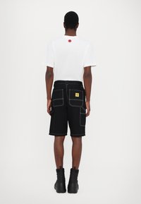 Black denim shorts with white stitching, two front pockets, and a rectangular yellow label. Paired with a plain white t-shirt.