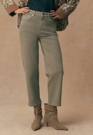 Modelis stāv, valkājot bēša krāsas augsta vidukļa džinsus, ziedu raksta jaku, melnu topu un taupe krāsas potīšu zābakus ar acu detaļām uz bēša fona.