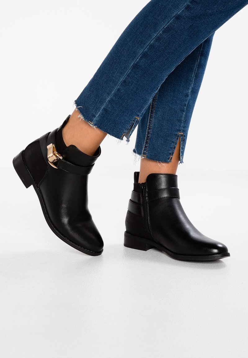 Mujer que lleva botas de cuero negras hasta el tobillo con detalle de hebilla dorada y jeans cortos azules con bordes deshilachados y aberturas laterales.