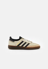 Beige sneakers met zwarte strepen, suède neuskap, witte veters en een bruine rubberen zool. Heeft gouden "Spezial" branding aan de zijkant.