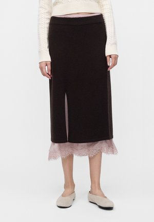 Vrouw draagt een zwarte midirok met een split aan de voorkant over een roze kanten slip, gecombineerd met een crèmekleurige trui en beige platte schoenen.