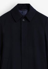 Abrigo de marinero con cuello clásico, cierre frontal de botones, dos bolsillos laterales y detalle de costura en contraste. Textura de tela suave y lisa.