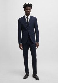 Marineblauer maßgeschneiderter Anzug mit einreihigem Sakko, slim-fit Hose und weißem Hemd. Eine schwarze Krawatte und polierte Schuhe runden den Look ab.