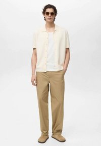 Camicia a maniche corte beige chiaro con bottoni sopra una canotta bianca, abbinata a pantaloni beige ampi e scarpe marroni, indossata da un uomo con occhiali da sole.