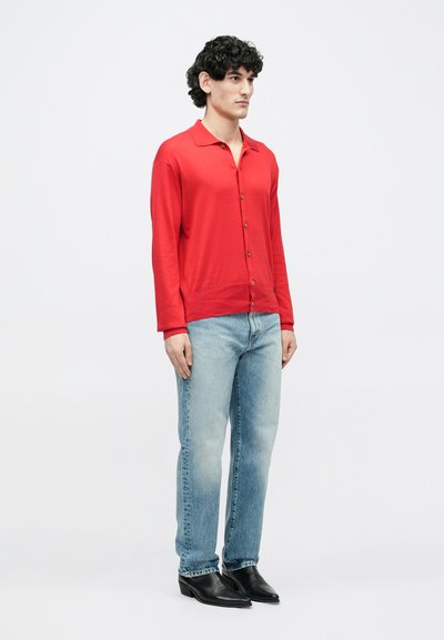 Fiatal férfi göndör hajjal, piros gombos kardigánt, világoskék farmert és fekete hegyes orrú csizmát viselve, egyszerű háttér előtt áll.