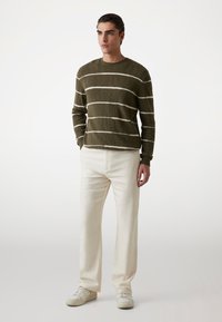 Olivgrüner gestreifter Pullover mit weißen Linien, Rundhalsausschnitt und gerippten Bündchen, kombiniert mit cremefarbenen, geraden Hosen und weißen Sneakers.