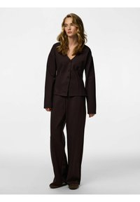 Hnědý dvoudílný outfit sestávající z halenky s výstřihem do V, zapínané na knoflíky, s dlouhými rukávy a přiléhavým střihem, a širokých kalhot ze stejného materiálu.