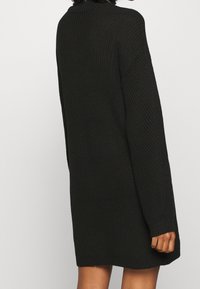 Svart stickad klänning med en strukturerad ribbdesign, långa ärmar och avslappnad passform. Synlig manschettdetalj och minimal design.