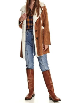 Brązowy płaszcz z sztucznego futra z białym podbiciem, dużymi guzikami, noszony na koszuli w kratę, zestawiony z jasnoniebieskimi dżinsami i brązowymi botkami sięgającymi do kolan.