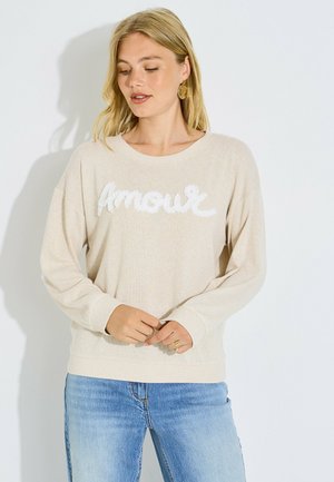 Mujer rubia con suéter beige con texto blanco "Amour" y vaqueros azul claro, de pie frente a un fondo blanco liso.