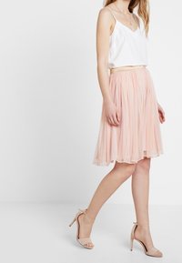 Nainen, jolla on päällään valkoinen hihaton toppi, vaaleanpunainen tyllymidimidi hame kultaisella vyötärönauhalla ja beige nilkkaremmit korkokengät, seisoo kyljellään.