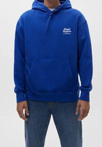 Blå hoodie med en framficka, justerbar dragsko-huva och vit broderad text på bröstet. Slät tyg och ribbade muddar.