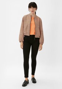 Gekürzte, karierte Bomberjacke in Orange und Weiß; kombiniert mit einem eng anliegenden orangefarbenen Oberteil, schwarzen Skinny-Jeans und schwarzen Loafers.