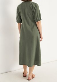 Oliwkowa sukienka do kolan o teksturowanej powierzchni, z krótkimi bufkami i luźnym krojem, w połączeniu z brązowymi płaskimi sandałami.
