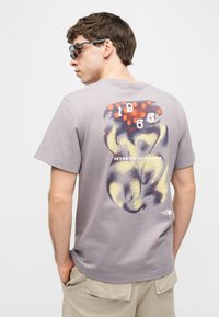 Άνδρας που φορά γκρι γραφικό T-shirt με αφηρημένο σχέδιο, αριθμούς "1966" και το κείμενο "Ποτέ μην σταματάς να εξερευνάς", συνδυασμένο με μπεζ σορτς και γυαλιά ηλίου.