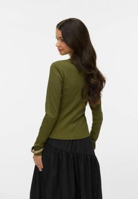 Žena s dugom tamnom kosom, odjevena u maslinasto zelenu majicu s dugim rukavima i crnu suknju, pozira djelomično okrenuta leđima i gleda u stranu.