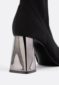 Črne elastične ankletske čevlje z sijočo, angularno kovinsko blok peto. Peta ima odsevno površino, kar dodaja vizualni kontrast.