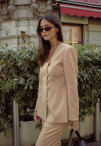 Beige tailored suit with a fitted blazer featuring round buttons. The texture is smooth, paired with black sunglasses and a black handbag.